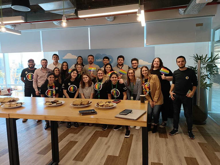 Personas en la oficina con camisetas con los colores de la bandera LGBTQ+, alrededor de una mesa con bocadillos.