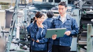 Two workers in a factory — a woman and a man, both wearing dark blue jumpsuits — look at plans on a tablet. / best field service management software