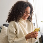 A smiling woman in a white sweater gets good customer service on her yellow mobile device from her couch