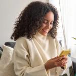 A smiling woman in a white sweater gets good customer service on her yellow mobile device from her couch