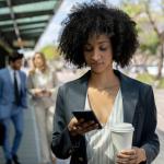 Black business woman at the bus stop holding her take out coffee and looking at her smartphone