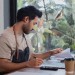 Attractive barista male or employee concentrate on stacks of tax declaration bills, restaurant owner using laptop for bookkeeping control payment, multitask small business lifestyle doing everything