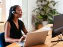 Woman wearing a headset, looking into a monitor and typing on a laptop, delivering customer service. / customer data in contact centers
