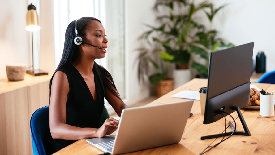Woman wearing a headset, looking into a monitor and typing on a laptop, delivering customer service. / customer data in contact centers