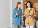 Two smiling women — one in a blue blazer and another in a yellow jacket holding a mobile table — discuss an upselling and cross-selling opportunity in a company’s reception area.
