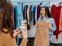 Two women shop for clothing in a store. One woman is taking a photo of her friend, who is holding up a light brown dress. / referral marketing