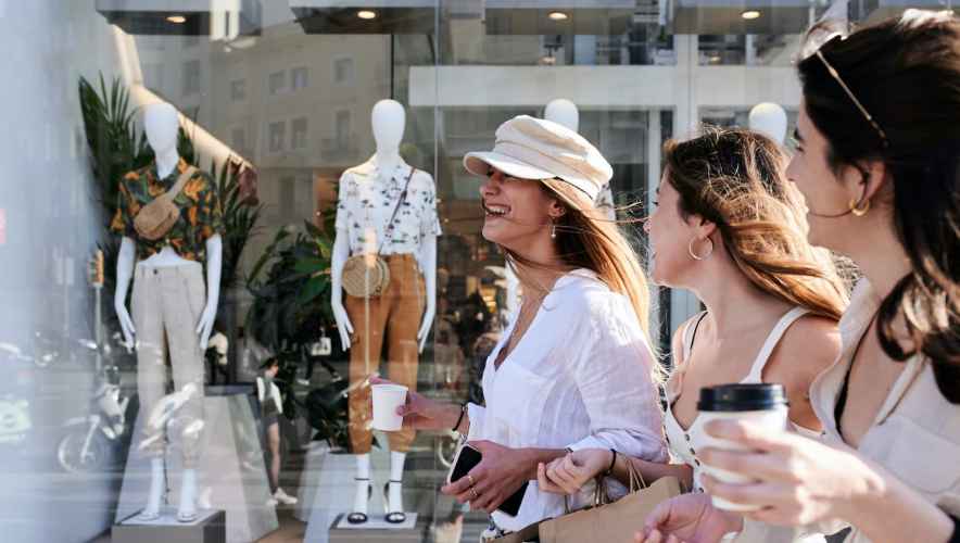 Young women drinking coffee walking past a clothing store window