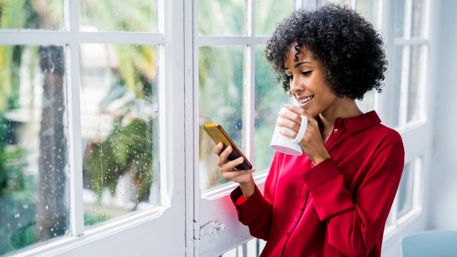 Woman in a red shirt checks her phone inside on a rainy day while holding a white coffee mug / email deliverability