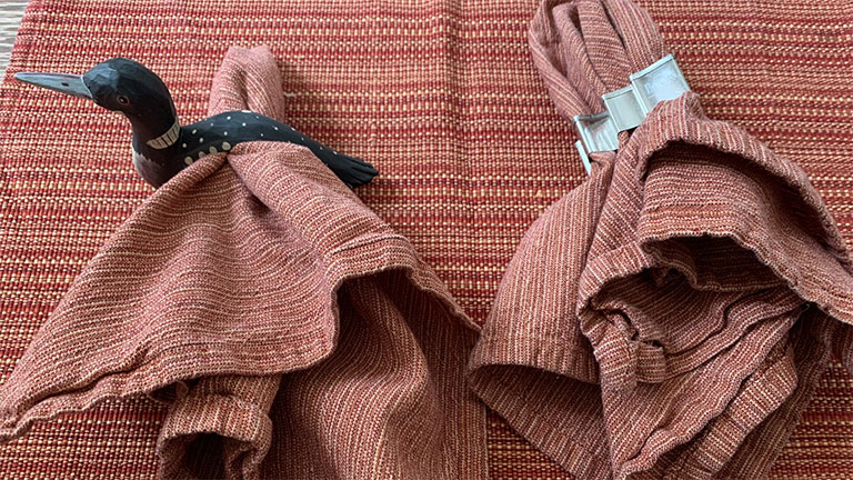 Two cloth napkins in rust-colored rings sit on a matching placemat, with one held by a decorative bird-shaped napkin holder.