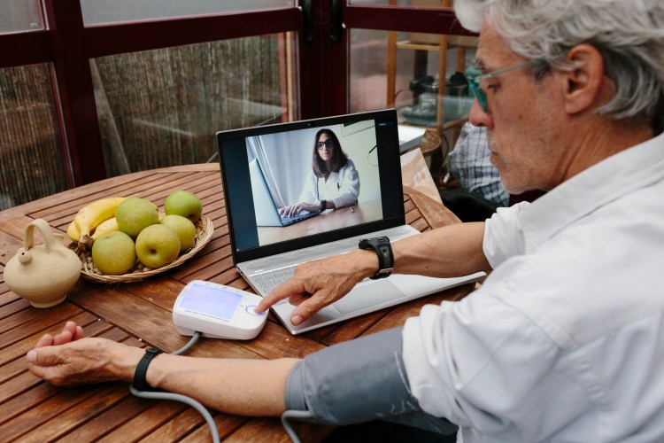 Elderly man checking his blood pressure at home in front of his laptop. He's having a video-chat with a female doctor who looks at him on the screen.