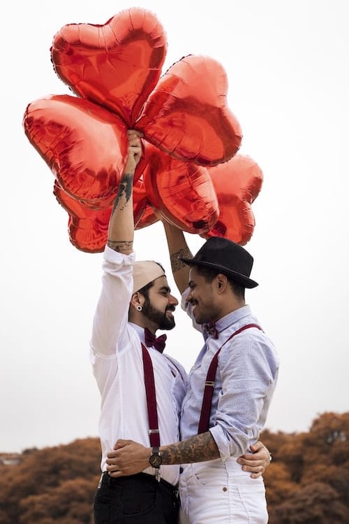 A photo of an LGBTQ couple looking lovingly into each others eyes, while holding heart balloons.
