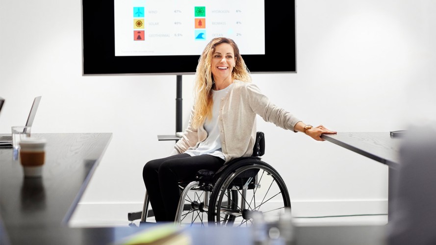 An employee in a wheelchair in front of a large monitor at work: NDEAM.