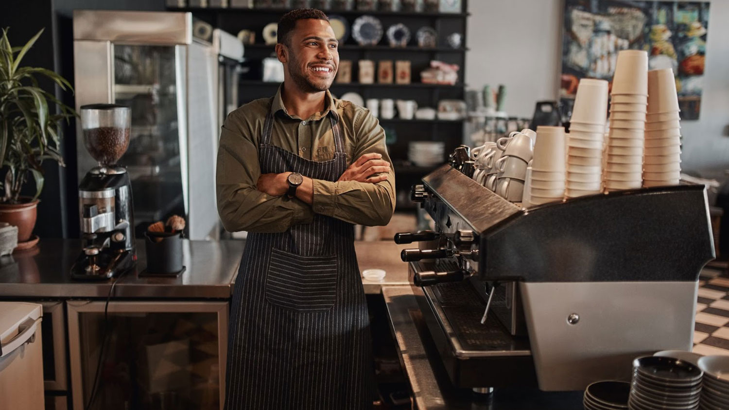 Black business owner in his coffee shop standing next to an espresso machine with his arms crossed.