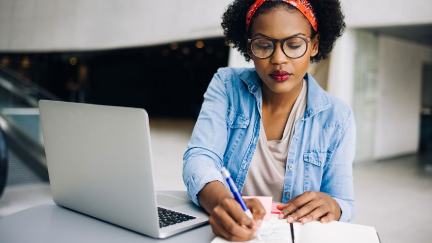 Black woman makes notes on paper while sitting in front of a laptop: small business pandemic entrepreneur