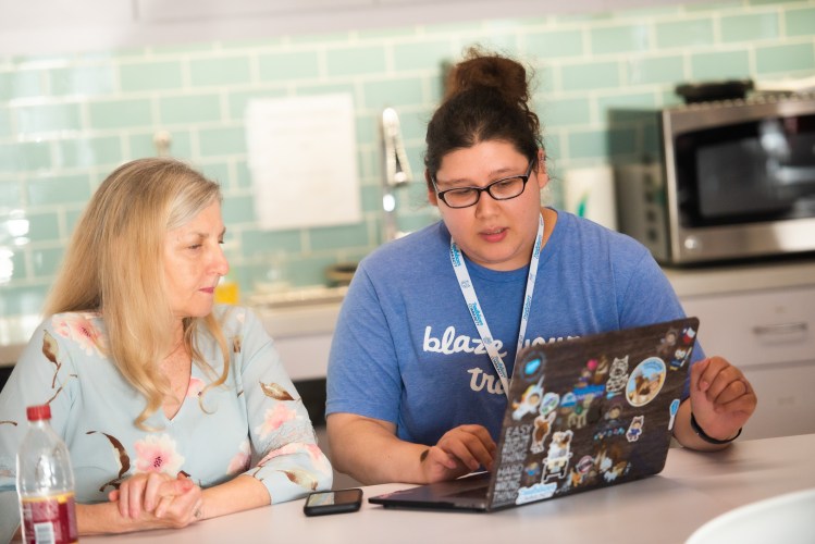 Two women looking at a laptop that has Salesforce stickers on the front of it. One woman is coaching the other.