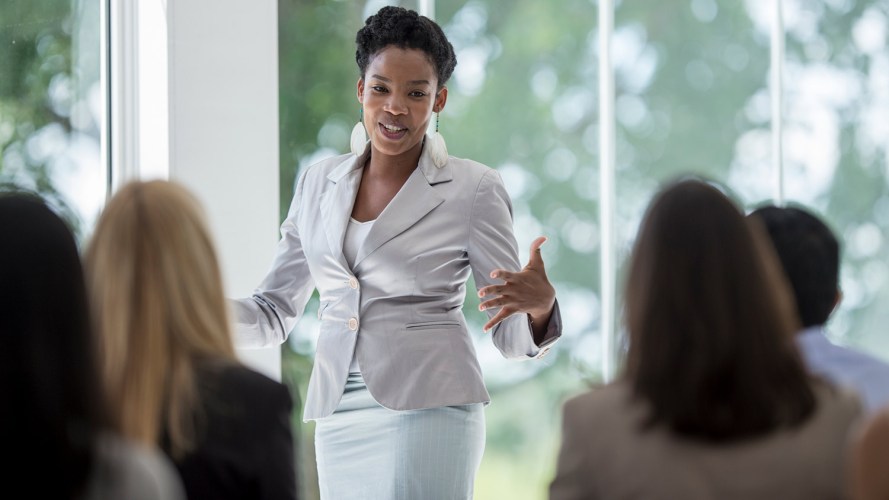 Black woman is white suit presenting in a conference room:women in leadership, women leaders, women and leadership