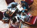 A group of five people sitting in a circle and working on devices such as phones, tablets, and laptops