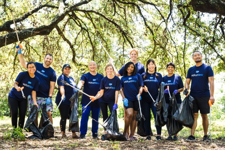 Salesforce Austin employees wearing blue "volunteer" t-shirts and holding trash bags and trash pickers under a forest canopy.