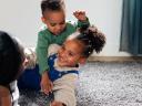 Two children play on a rug while smiling at a caregiver.