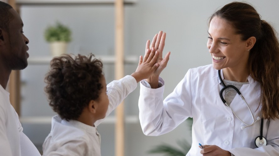A parent holds their child while the child high-fives a doctor