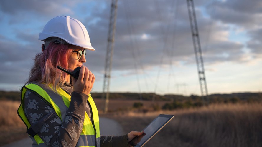 A woman wearing a hard hat views telecom innovations on a tablet near a cell phone tower