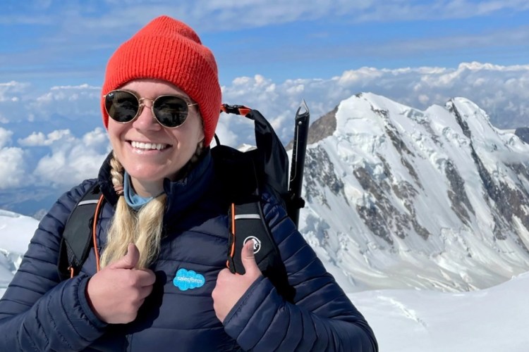 Florine smiling, she is wearing a jacket with the Salesforce logo while climbing. There is a snowy mountain in the background