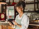 Woman smiling in a bakery, wearing a green apron, holding a tablet and learning more about how automation from the communications industry can help her small business.