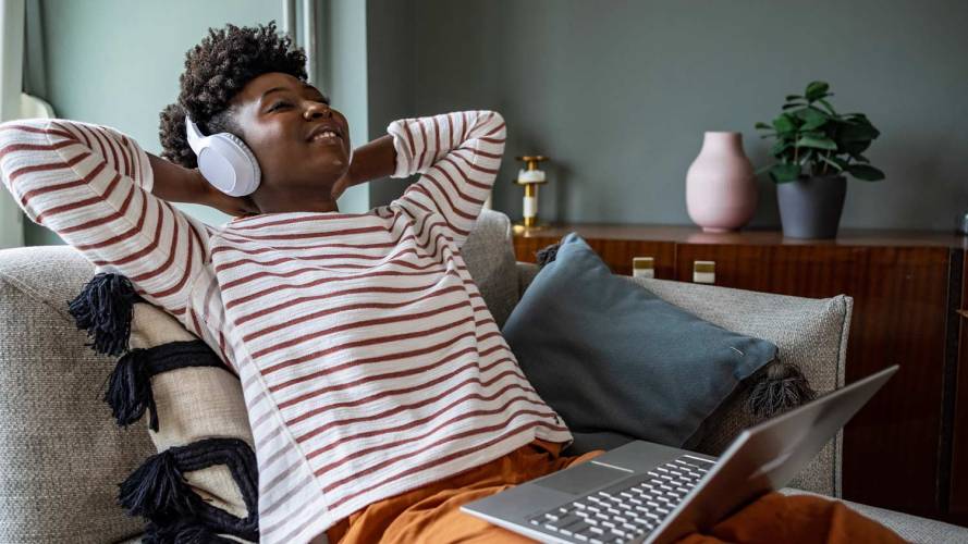 Woman sitting on her couch, wearing a striped shirt and listening to music on her headphones with a laptop on her lap / subscriber relationships