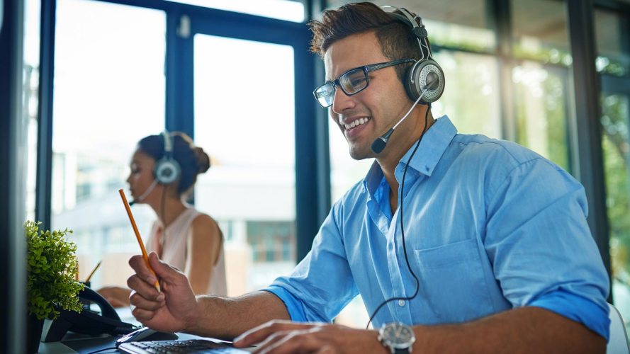 Male service agent in a blue shirt, working from a contact center, smiling while talking with a customer on his headset / invest in customer support