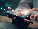 Image shows a pair of hands holding and tapping a mobile phone. There are many heart emoji floating above the screen.