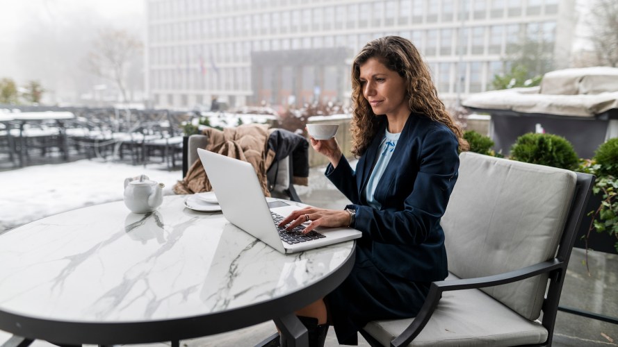 Businesswoman in a blue jacket, sitting at a table outdoors during the winter, checking her laptop while drinking coffee out of a white mug / sandbox preview