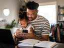 Father sitting at a desk at home, holding his toddler daughter while he checks his phone / customer experience