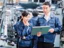Two workers in a factory — a woman and a man, both wearing dark blue jumpsuits — look at plans on a tablet. / best field service management software