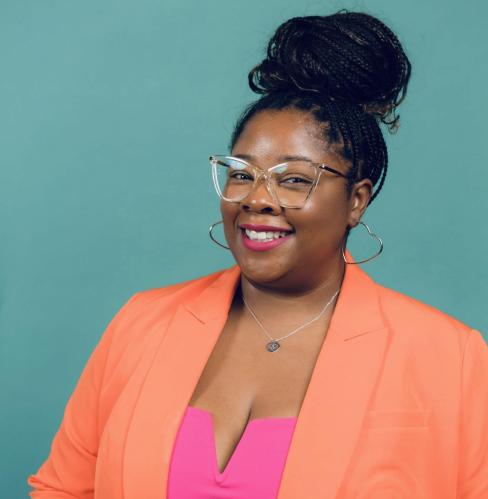 Photo of Kim wearing an orange blazer with a green background behind her.
