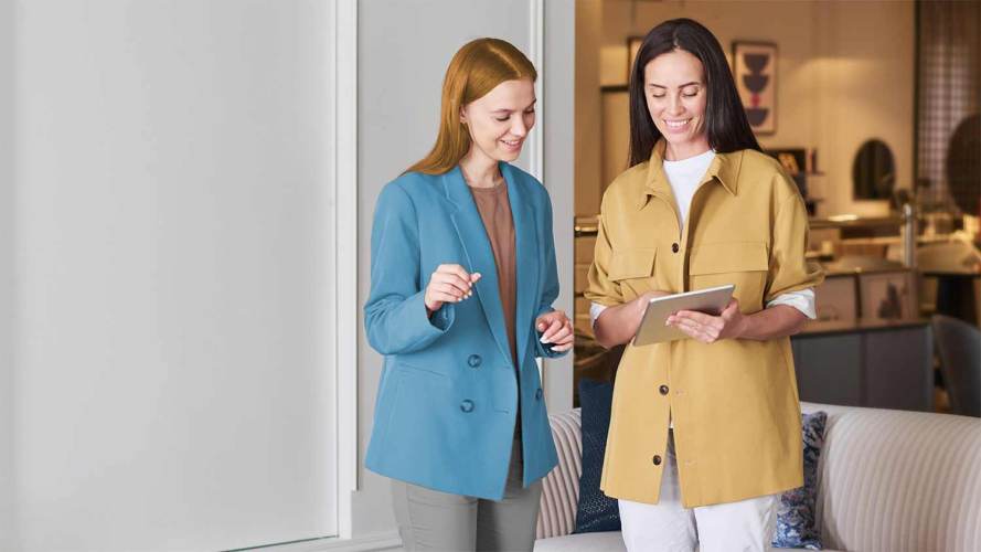 Two smiling women — one in a blue blazer and another in a yellow jacket holding a mobile table — discuss an upselling opportunity in a company’s reception area.