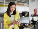Female office worker, professionally dressed in a bright yellow jacket, stands near her desk while she checks her employee self-service system from her smartphone