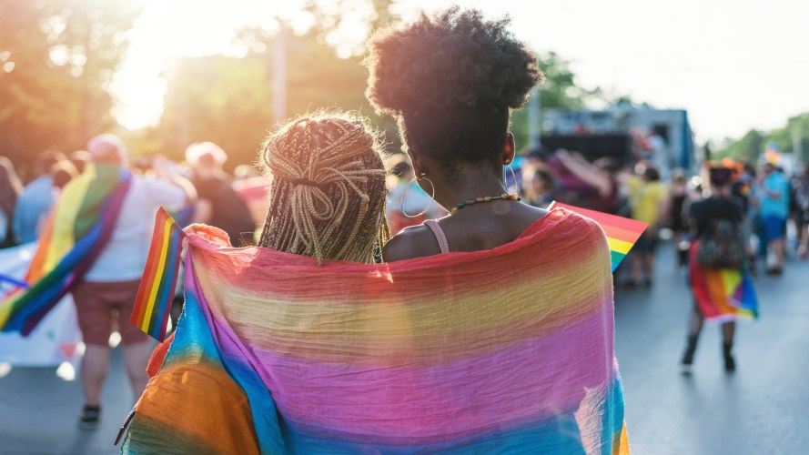 Rear view image of young couple walking with the pride event, hugging and waving pride flags during transgender awareness week