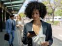 Black business woman at the bus stop holding her take out coffee and looking at her smartphone