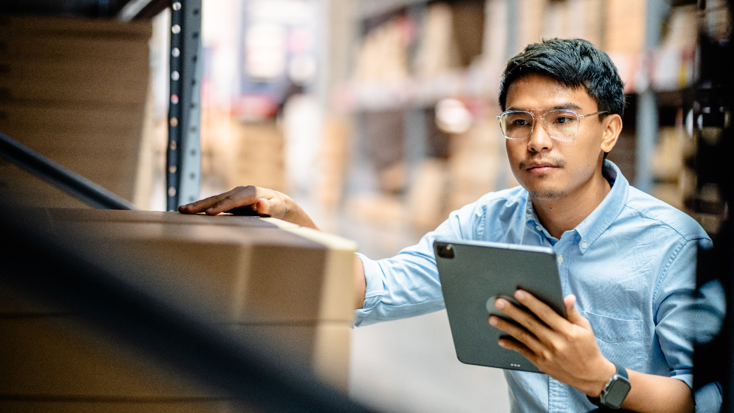 A man wearing glasses and a blue collared shirt holds a tablet as he looks at inventory in a warehouse.