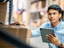 A man wearing glasses and a blue collared shirt holds a tablet as he looks at inventory in a warehouse.