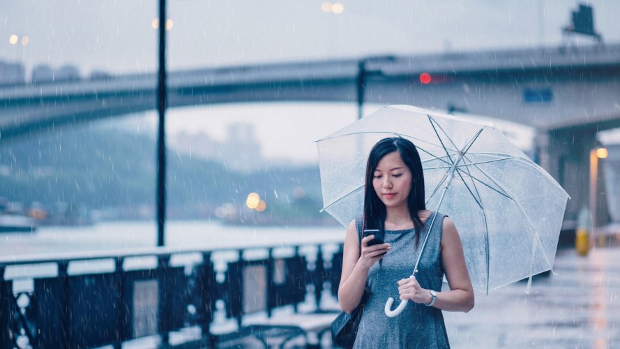 Photograph of a young businesswoman, dressed in a gray dress and holding a clear umbrella, checking her phone on a rainy, overcast day. / drip marketing