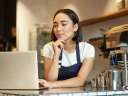 A young woman, working in her coffee shop and dressed in a white shirt and a dark blue apron, checks her laptop on the counter. / email marketing for small business