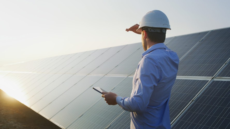 A mobile worker stands next to solar panels, shielding his eyes while looking toward the sun and a bright future.