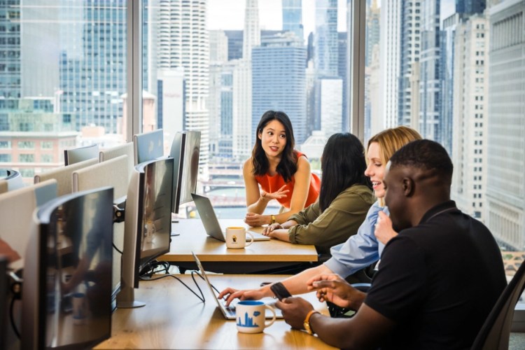 A diverse group of professionals is collaborating and conversing in a modern office with large windows.