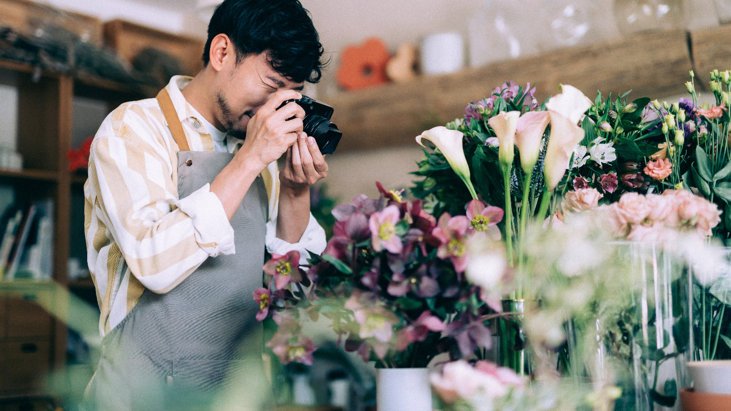 Male employee in a flower shop, wearing a pink stress shirt and an apron, takes a photo of some flowers in his store / digital marketing for small business