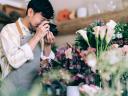 Male employee in a flower shop, wearing a pink stress shirt and an apron, takes a photo of some flowers in his store / digital marketing for small business