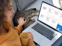 Image of a woman petting a cat and holding a mug while looking at a customer portal on her laptop.