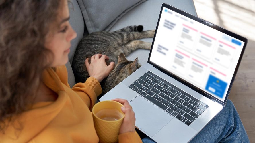 Image of a woman petting a cat and holding a mug while looking at a customer portal on her laptop.