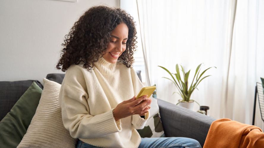 A smiling woman in a white sweater gets good customer service on her yellow mobile device from her couch