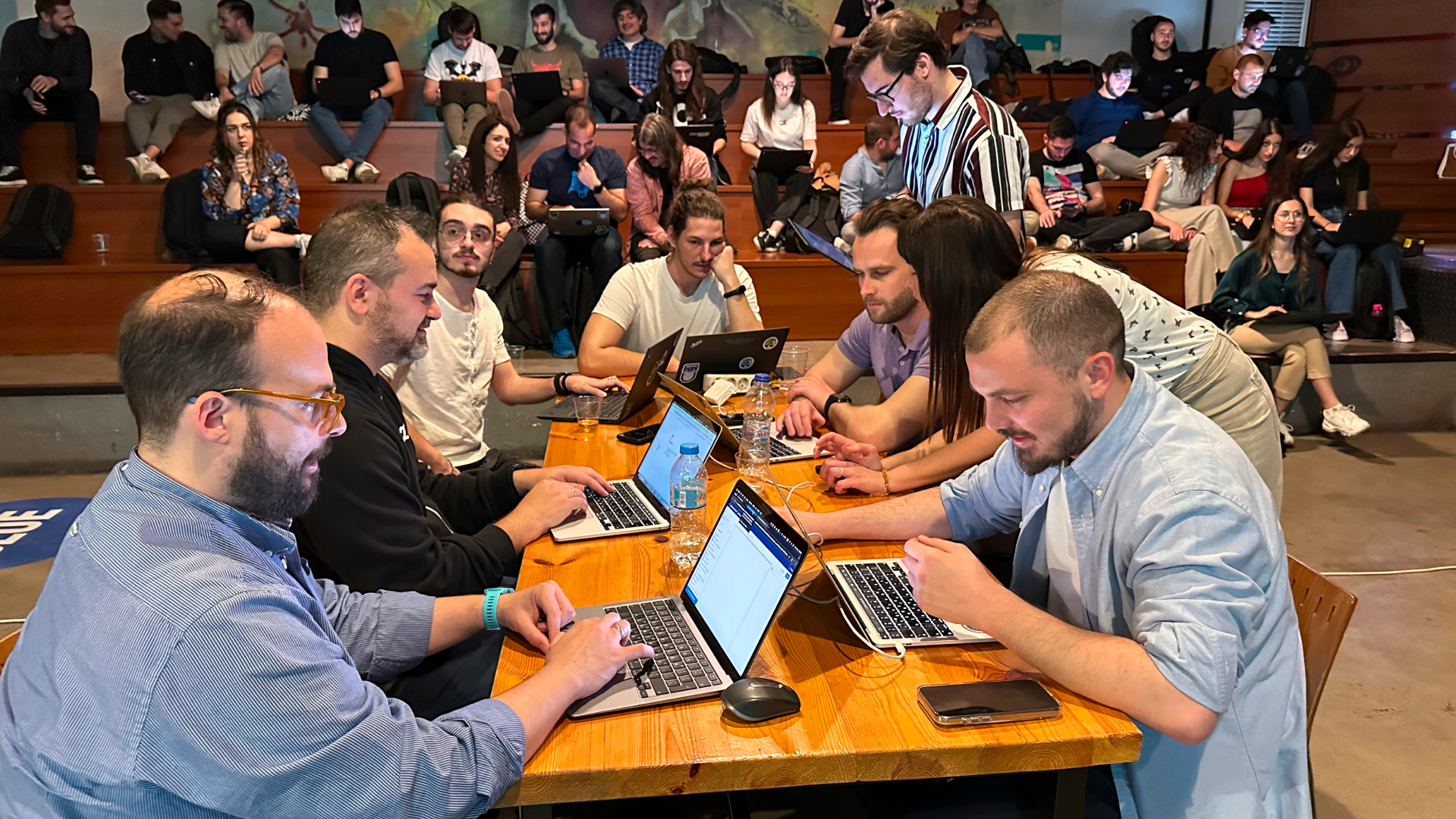Seven people at one table on laptops at the AI + Data Community Tour stop in Greece, with others working in stadium seating in the background.